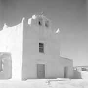 The front of the Church of San Jose at the Laguna Pueblo in New Mexico has a large bell tower with two bells and a large cross. A large window is centered over the large, wooden door; a smaller entrance is in the adjacent one-story section of the church. The adobe surface is cracked in several areas, particularly above the main door and window.  The church is dated at approximately 1699.