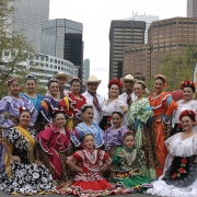 Men, women, girls and teenagers, members of Jeanette Trujillo-Lucero's Fiesta Colorado Dance Company pose on Broadway during Cinco de Mayo celebrations in Denver, Colorado. The girls and women wear traditional Mexican style dance costumes in bright colors decorated with lace and ribbon. They all have their hair pulled up with flower decorations. The men wear white shirts, bandannas and straw hats. Booths and high rise buildings are in the distance.