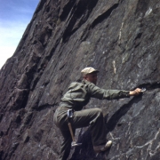 Tenth Mountain Division soldier, Joe Stettner, climbs a steep rock face near Camp Hale, Colorado. He wears a cap, khaki uniform, and soft-soled shoes. Hammer and carabiners hang from his belt as he clips a carabiner to a piton.