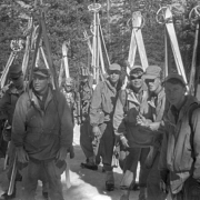 Shows a group of Tenth Mountain Division skitroopers standing on the snow in the mountains. They have their skis and poles attached to the sides of their knapsacks. All of the are wearing caps, anoracks, pants and boots with gaiters. Four of them are also wearing ski goggles.