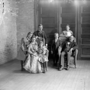 Group studio portrait of Adolph Joseph Zang Family; seated, left to right:  mother (Wilhelmina "Minnie" Louise Vogt) and youngest daughter (Louise) sharing chair, father (Adolph Joseph Zang); standing, left to right:  middle daughter (Minnie Elizabeth), eldest son (Philip Adolph II), youngest son (Adolph Frank), and eldest daughter (Gertrude).  Two chairs are wicker; background is bricked corner at left, three doors center and right; wood plank flooring.