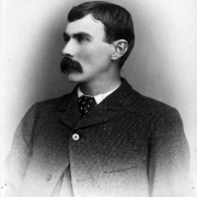 Head and shoulders studio portrait of Ed Creighton, Gunnison, Colorado; he wears a tweed wool sack coat with narrow lapels with top button fastened and a cravat. He has short combed hair and a heavy mustache.