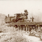 View of a Denver and Rio Grande Railroad train at Maysville (Chaffee County), Colorado.  Shows locomotive number 52 and box cars on the bridge at the North Fork of the South Arkansas River. Men in work clothes stand on and near the train, snow covered mountains are in the distance.