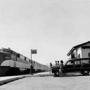 View at the Southern Pacific Railroad depot in Palm Springs (Riverside County), California. Women pose by a 1948 Ford "woody" station wagon and the Mission Style building; the Southern Pacific locomotive and streamliner "Golden State" waits by a sign that reads: "Palm Springs Stage."