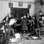 Interior view of the Cooper home in Idaho Springs, Colorado, shows six musicians playing their instruments. Included are: coronet, valve trombone, piano, clarinet, and two violins. The upright piano has a decorative wooden engraving on the facing board and a star patterned cloth draped over it. A doll on top of the piano is leaning against a basket of flowers.