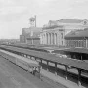 View of the Union Pacific "City of Denver" and the Chicago, Burlington & Quincy Railroad "Denver Zephyr" trains, umbrella sheds and passenger platforms at Union Station in Denver, Colorado. The railroad depot is a rusticated stone building. The 20th (Twentieth) Street viaduct is in the distance.