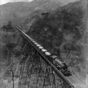 View of a steel Bingham & Garfield Railroad trestle over Markham Gulch in Bingham Canyon, Salt Lake County, Utah. A Bingham & Garfield 0-8-8-0 locomotive number "101" hauls copper ore on the trestle. Shows wooden Utah Copper Company (later Kennecott Copper Corporation) buildings, probably tramway cables and a tower. The Oquirrh Mountains are in the distance.