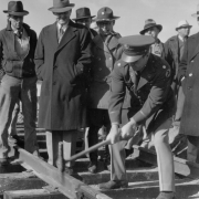 View of a dedication ceremony with men in military uniforms, or suits and overcoats, at Lowry Army Air Force Base in the Lowry Field neighborhood of Denver, Colorado. The men watch an officer hammer a spike into railroad tracks constructed by the Work Projects Administration (WPA)  The men wear caps and fedoras. The tracks were constructed to connect Lowry to the Union Pacific Railroad line.