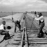 Work Projects Administration (WPA) railroad construction workers in overalls and fedoras use a hammers and a crowbar to install rails at a switch on railroad tracks near Lowry Air Force Base in the Lowry Field neighborhood of Denver, Colorado. Shows a railroad signal and a culvert beside the tracks. The tracks were constructed to connect Lowry to the Union Pacific Railroad line.
