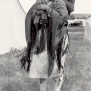A Native American (Arapahoe) woman smiles and poses in Fort Washakie (Fremont County), Wyoming, with her baby, carried on her back. She wears a blanket with long fringe, a gingham dress, and high top leather moccasins with applique. A car is in the distance.