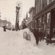 Snow fills the street of a town; people pose by shop windows, snow shoes, and a mortar and pestle. Business signs read: "Suomalainen Aptecki" (Finnish pharmacy), "Dr. G. J. Sorsen," "A. B. Turner, Bankers," "Star Clothing House," and "Hoskings Co. Dry Goods." Utility poles by the street have multiple insulator crossbeams.