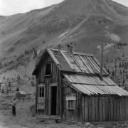 An unidentified woman stands at the side of a large, two-story log cabin in Chihuahua Gulch near Montezuma in Summit County, Colorado. A dirt road runs from the foreground past the cabin. Pine trees stand at the base of a mountain ridge in the background.