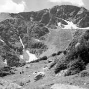 A group of three unidentified people is in the left foreground in the Mammoth Basin west of Apex in Gilpin County, Colorado. One person sits alone and the other two are farther away, one sitting on a rock, the other standing. In the distance, the rocky ridge of James Peak has small patches of snow on it. James Peak stands where Gilpin, Grand and Clear Creek Counties converge in Colorado.