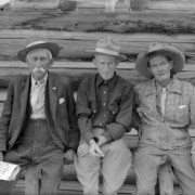 John A. (Jack) Gilfillan, John H. Kemp (father of the photographer) and J. B. Rowley sit on a log railing in front of a log cabin  in Eldora, a mining town in Boulder County, Colorado. The three men each helped found the town in the late 1890s. All three wear hats. Gilfillan and  Rowley hold newspapers in their hands, and Kemp has a cigarette in his right hand.