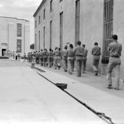 Convicts march with arms crossed at the State Penitentiary in Canon City, Colorado. The concrete wall next to them has barred windows. The inmates approach cell house #1, a three-story concrete building with barred windows.