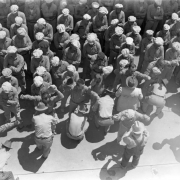 Convicts in golf hats are frisked by guards in cowboy hats at the State Penitentiary, in Canon City, Colorado. Prisoners waiting their turn stand with arms crossed.