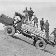 View of a Coleman flatbed truck on an incline with the city of Denver, Colorado, in the distance. A group of well-dressed men and workers ride on the truck loaded with sacks of probably sand. The surrounding landscape is barren. The state capitol building is visible in the distance.