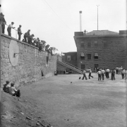 Convicts play basketball at the State Penitentiary in Canon City, Colorado. An armed guard watches from the metal roofed admissions building, with its anemometer. Hoops and backboards are mounted on the rock wall, and prisoners sit on top and lean on the railing. Stairs connect the two levels. Men sit on a dirt pile in the foreground.  Metal smokestack is in the background.