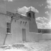 View from the side of the adobe St. Francis of Assisi church in Ranchos de Taos (Taos County), New Mexico. Shows an adobe wall with a doorway and window, wooden vigas, and two bell towers with crosses on top.