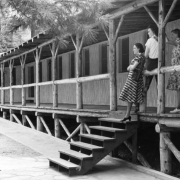 Teenage girls pose at the Beulah resort near Pueblo, Colorado, on the log railing of an open porch fronting a frame, one-story dormitory. The walks at the bottom of the steps are concrete with curved 'grout' lines.