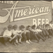 Young men, probably Devlin's Zouaves with the Sells Floto Circus and Buffalo Bill's Wild West Show, pose seated on the ground in Great Falls (Cascade County), Montana. The young men are near a billboard that reads: "Drink American Beer." The boys wear caps, shirts and pants. A circus trailer is seen in the background.