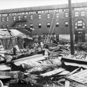 Flood debris in Pueblo, Colorado, includes carved wood molding, stamped ceiling metal, door, crates, wheels, lumber, and mud. A frame building has roof damage and a sign: "Mammoth Corral." A shed lays sideways in front of a four-story brick warehouse with arched windows and signs: "The Colorado Bedding Co. Established 1894." White residue covers the first  floor, showing the peak of the high water.