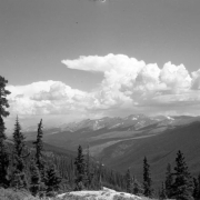Pine trees stand on a hillside in the foreground in This view looking north toward a ridge of mountains in the distance from Berthoud Pass on the border of Clear Creek and Grand counties in Colorado. Small patches of snow are on the mountains in the distance. Cumulus clouds form a thunderhead anvil in the distance.