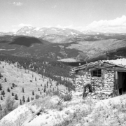 An unidentified man leans over, possibly to tie his shoe, in front of stone cabin in the foreground in This view near Jenny Lind Gulch, a tributary of South Boulder Creek near Apex in Gilpin County, Colorado. Pine trees line the rolling hills leading back to a ridge of mountains in the distance.