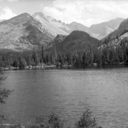 Rugged mountains stand in the distance in This view of Bear Lake in Rocky Mountain National Park in Larimer County, Colorado. Tall pine trees stand on the far shore of the lake.