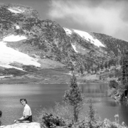 Two unidentified women sit on rocks in the left foreground on the  near shore of St. Mary's Lake looking toward St. Mary's Glacier in Clear Creek County, Colorado. Tall trees stand on the shore of the lake in the right midground and near where the two women sit.