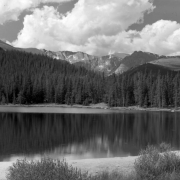 View of Echo Lake near Mount Evans in Clear Creek County, Colorado; pine forest and narrow beaches surround the water. Cumulus clouds top snow dotted mountain peaks in the distance.
