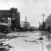 Water, mud and debris fill Union Avenue in Pueblo, Colorado. Brick storefronts show signs: "Klein & Leiser," "Colonial Jewelry," "Exchange Rooming House," "Weisel Seed," "Star House," and "Economy Clothing." Cast iron street lamps with milk glass globes line the thoroughfare. The Board of Trade Building, City Hall, and the turret of the Riverside Block show above other structures. The steel arch of the Arkansas River bridge is past wrecked automobiles and piled lumber.