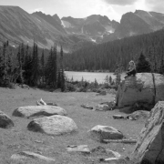 An unidentified woman sits on a large boulder near Long Lake west  of Ward in Boulder County, Colorado. Additional smaller boulders are also in the foreground. Tall pine trees stand along the shore of the lake in the  background. A ridge of mountains is in the distance.