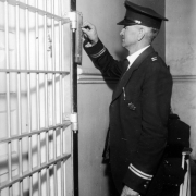 Holding a ring of keys and a cigar, This uniformed Captain at the State Penitentiary in Canon City, Colorado, operates a locking device next to hinged prison bars. A padded chair is in the background.