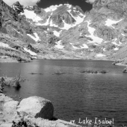 A large rock and tree branches stand in the foreground on the shore of Lake Isabelle in Boulder County, Colorado. Several smaller rocks also lie in the foreground. A ridge of jagged mountains with patches of snow on them is in the distance on the far shore of the lake.