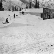 Snow is piled up in front of the Assay Office of Welch and Bancroft on Eldorado Avenue in Eldora, a mining town in Boulder County, Colorado. An unidentified man sts on a log in the left midground at the base of a mound of snow where two young boys play. One of the boys holds a sled propped up on one end. The other stands above him on the pile, possibly wearing skis.