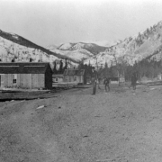An unidentified man walks across a dirt street in Eldora, a mining town in Boulder County, Colorado. The town was originally named Eldorado but its name was changed to avoid its being confused with Eldorado, California. Two children walk behind the man. A small group of unidentified people stands near a log cabin in the midground. Additional cabins line the dirt street. A ridge of mountains is in the distance.