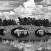 Arching bridge over Lake Clara at Mineral Palace Park in Pueblo, Colorado, is of random, pale stone with dark grout. Arches underneath provide support, and all is reflected in the rippled water in the foreground.
