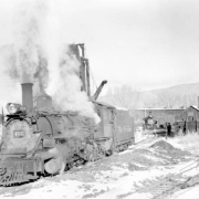Three-quarter view of left side of engine, from front end, close view; at water tank; smoke, steam, snow; shops in background. Photographed:  Ridgway, Colorado, November 18, 1951.