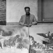Ciriaco Arellano, a Mexican immigrant to Colorado and self tough artist, poses with two of his paintings in probably Denver, Colorado. The painting on the left shows a herd of sheep caught in a blizzard, the one on the right is the Mexican national flag. It shows an eagle with a serpent in its talons, it stands on a cluster of banners over a stylized string of prickly pear cactus.