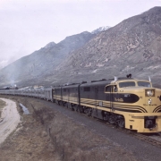 View of the eastbound California Zephyr, pulled by Denver & Rio Grande Western locomotive #601, in the Salt Lake Valley, Salt Lake County, Utah. Shows the train, with domed observation cars, near the Wasatch Mountains. The KF Steel plant is in the distance.