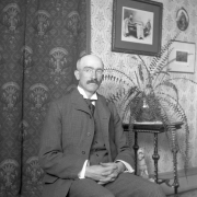 H.H. Lake poses for a portrait in a suit with a polka-dotted tie, possibly at his home in Central City, Colorado, where he worked at the  First National Bank for 47 years. He wears small glasses and has a large moustache. A fern stands on the table in the background. Several pictures hang on the wall behind him. Lake holds his hands folded in his lap.