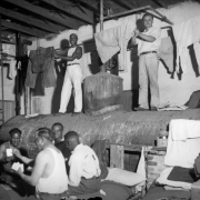 Black and white convicts at the State Penitentiary in Canon City, Colorado, work in the prison laundry. The interior ceiling is open- beamed, and a riveted metal tank below has pipes, valves and a gauge;  men stand on it, hanging clothes to dry. Others sit on a cot below playing a card game.