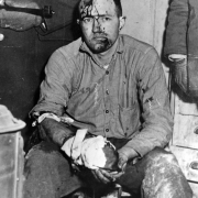 Heilman, recaptured after the 1947 escape from the State Penitentiary in Canon City, Colorado, has a rifle aimed at his blood spattered, half shaved head. His hand, wounded by gunshot, is wrapped in bloody rags; he sits holding it. The guard holding the gun wears mittens. A trash can and dresser flank the convict. Heilman was convicted of kidnapping Deputy District Attorney Fred Pferdesteller in Denver, 4/24/45.