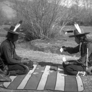 Antoine Moiese ("Grizzly Door") and Michael, two Native American men on the Flathead Indian Reservation in western Montana, sit on a striped blanket playing cards. Both men wear wide-brimmed hats with single feathers stuck in their bands. Both also wear vests and beaded necklaces. The man on the right has several ermine fur tassles hanging from his vest. A small creek is in the background with several shrubs along its bank.