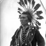 Chief Eagle, a Native American man on the Flathead Indian Reservation in western Montana, poses wearing a warrior's headdress in front of a teepee on the reservation. He wears large, round earrings, numerous beaded necklaces hanging around his neck, an otter sash across his chest, and holds a long spear, possibly a coup-stick, also wrapped in fur.