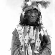 Francois Dead Horse, a Native American man on the Flathead Indian  Reservation in western Montana, poses in front of a teepee on the reservation. He wears a fur hat adorned with feathers, a buckskin shirt with beads sewn on it, and ermine fur tassles adorning it.