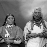 Studio portrait of Grizzly Bear and Marie, two Native Americans (Salish) on the Flathead Indian Reservation in western Montana. They pose in front of a painted canvas drop. Grizzly Bear holds a ceremonial pipe and wears a woven fringed tunic and has his long hair in braids, he has a scarf tied around his neck. Marie wears a polka-dotted dress and also holds a ceremonial pipe. She has several necklaces with an eagle feather hanging around her neck. Her hair is in two thick braids.