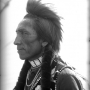 Louis Charlemain, a Native American man on the Flathead Indian Reservation in western Montana, poses for a formal, side-angle, bust portrait. He wears a fur roach on the top of his head. Strips of fur are wrapped around his long braids. He has an earring in his left ear and wears several beaded necklaces. His vest has beaded flowers decorating it. Charlemain also has markings under his eyes, possibly tattoos or paint. He is also known as Louie Molman.