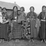 Four Native American women, including Louise Kumkumpoo Qumqmtpu, Agate Ogden Finley, Mrs. Latati and Agnes Incashola, pose with two small children on the Flathead Indian Reservation in western Montana. The women all wear their hair in braids. Their dresses are made from floral and checkered patterned materials. Each of them also wears several necklaces around her neck. Three of the women hold patterned, wool blankets. A long, wood building is in the left background.